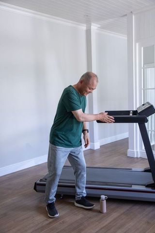Senior asian man checking smartwatch while leaning on treadmill in bright home gym