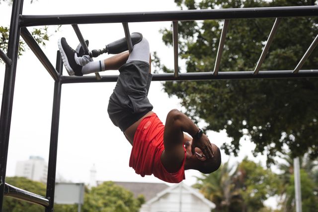 This image shows a disabled man with a prosthetic leg performing crunches while hanging from monkey bars in an outdoor gym. It highlights themes of fitness, determination, and healthy lifestyle. This image can be used for promoting adaptive sports, fitness programs for people with disabilities, and motivational content.
