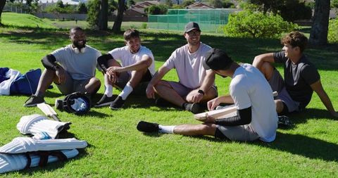 Diverse male friends sitting on grass chatting after cricket, casual outdoor team bonding