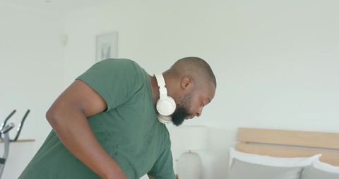 African american man making bed in modern minimalist bedroom wearing headphones