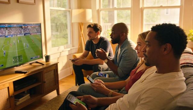 Friends gaming together on sofa watching soccer match on tv with controllers and phones