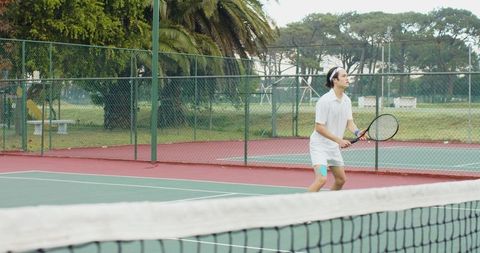 Asian man playing tennis on outdoor public court wearing white outfit and teal knee tape