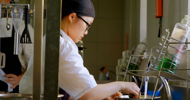 A chef is carefully preparing a meal in a busy commercial kitchen, demonstrating focus and professionalism. The chef is wearing a black cap, white uniform, and glasses, emphasizing her commitment to food preparation. This image can be used for culinary-themed articles, restaurant marketing, cooking school promotions, and other food-related content, showcasing dedication and skill in professional cooking.