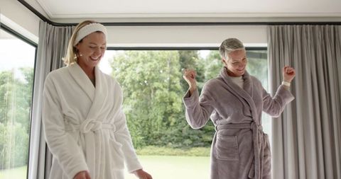 Two women wearing plush robes laughing by glass doors in bright modern home