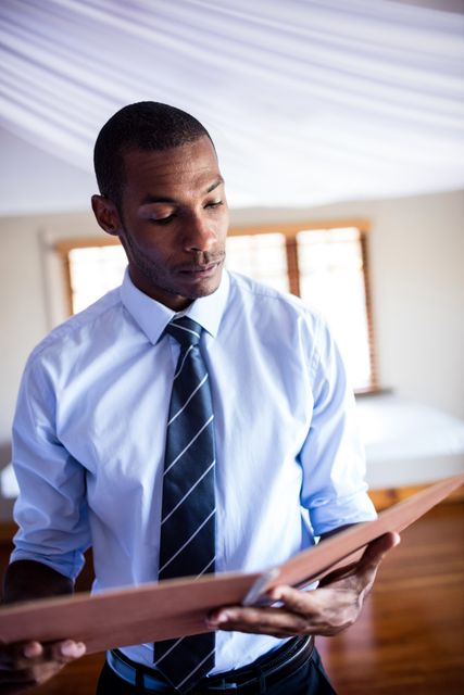 This image depicts a male supervisor in a hotel room, attentively reviewing a file. He is dressed in professional business attire, including a shirt and tie, indicating a formal work environment. This image can be used for articles or advertisements related to hotel management, corporate training, business professionalism, or the hospitality industry. It is suitable for illustrating concepts of attention to detail, professionalism, and managerial responsibilities.