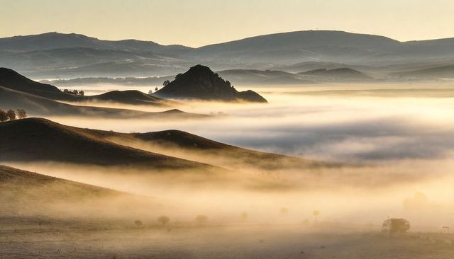 Golden sunlight bathing mist-filled valley with layered hills, rocky outcrop and lone vehicle
