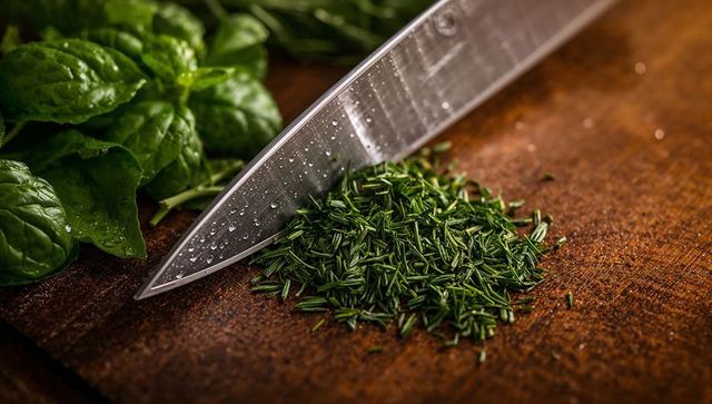 Chef knife resting on rustic cutting board with chopped rosemary and fresh greens closeup
