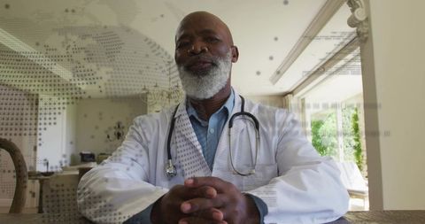 Senior physician consulting from home, clasping hands, wearing white coat and stethoscope