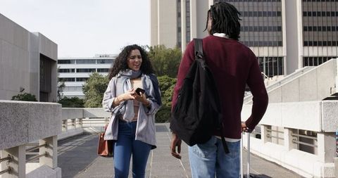 Diverse coworkers chatting on walkway, woman holding smartphone, man pulling suitcase