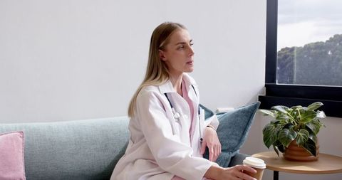 Female clinician relaxing on clinic sofa holding coffee wearing lab coat and stethoscope