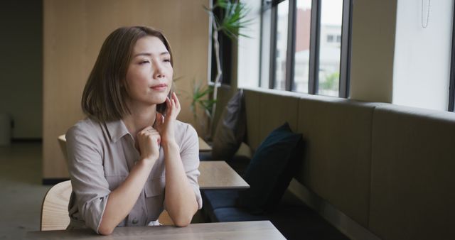 Image depicts a woman sitting alone in a modern cafe by large windows, looking contemplative. Suitable for use in stories about reflection, relaxation in urban environment, or modern lifestyle blogs, advertisements for cafes and coffee shops, as well as articles discussing personal growth and introspection.
