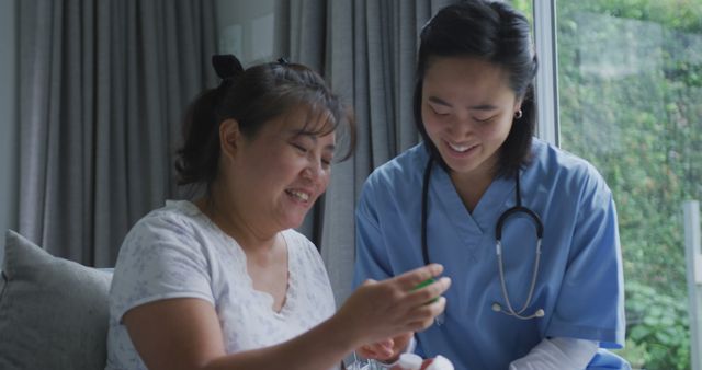 An elderly Asian woman is sitting indoors in a cozy home environment with a smiling nurse or caregiver assisting her with medication. The nurse is wearing blue scrubs and has a stethoscope around her neck, indicating her professional role. This image is excellent for illustrating themes of home healthcare, elderly caregiving, nursing support, and medical assistance. It can be used in brochures, websites, or advertisements related to healthcare services, senior care, and medical professional training.