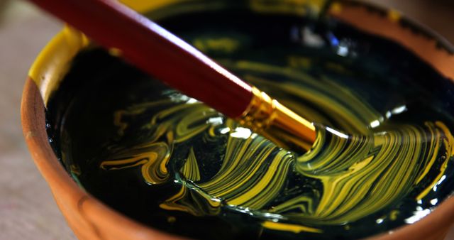 Visual closely captures a paintbrush mixing yellow and black paint in pot, showing detailed swirls of color. Ideal for usage in art materials promotions, creative tutorials, or articles about painting techniques.