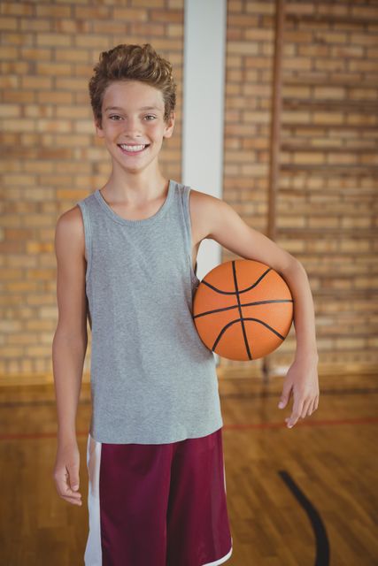High school boy smiling and holding a basketball in a gym with a brick wall background. Ideal for use in educational materials, sports promotions, youth fitness campaigns, and articles about teenage athletes.