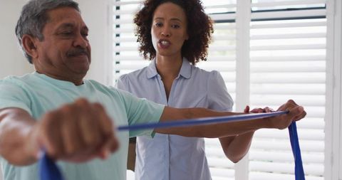 Senior man performing resistance band exercises while caregiver guiding home rehabilitation