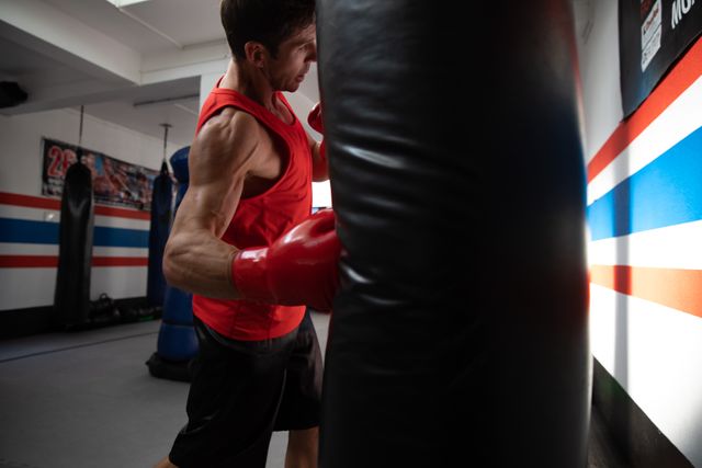Side view of a young biracial male boxer with short dark hair, wearing red tank top, exercising in a boxing gym, punching a punchbag.