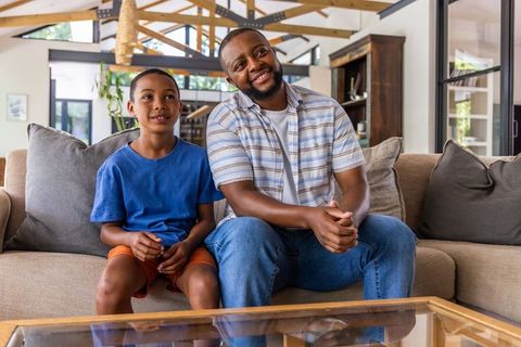 African american father and son sitting on modern sofa smiling, warm home family bonding