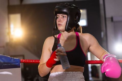 Female boxer resting in ring corner wearing headgear and pink glove holding water bottle