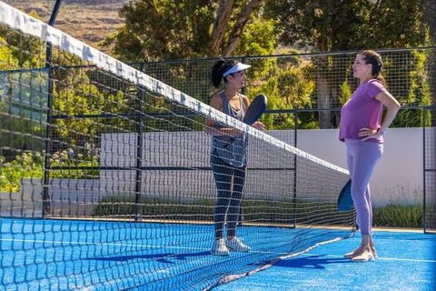 Mature women chatting at net on blue outdoor pickleball court holding paddles with visors
