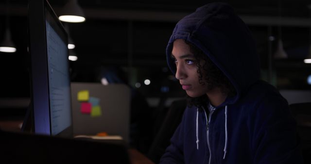Individual wearing blue hoodie intently working at computer in dark office setting. Sticky notes visible on nearby desk, indicating ongoing tasks. Ideal for themes involving cybersecurity, tech industry, night shifts, focus, and dedication.