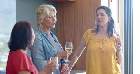Middle-aged women chatting over wine at home bar in bright modern kitchen