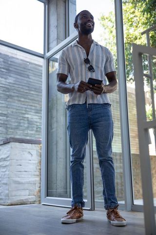 Casual african american man standing in modern glass lobby holding smartphone and smiling