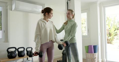 Female personal trainer coaching client lifting dumbbells in bright home gym session