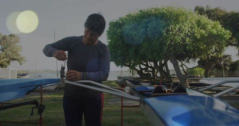 Adjusting rigger on blue rowing shell by experienced woman at waterfront boatyard
