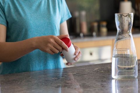 African american woman opening red-lid supplement container on kitchen counter with carafe
