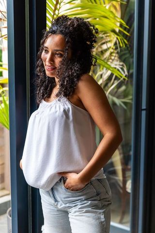 Curly-haired african woman leaning by glass door with tropical plants, casual summer look
