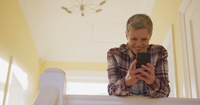 Mature woman standing in a well-lit home, smiling and laughing while texting on her smartphone. Ideal for advertising communications technology, promoting positive vibes or showcasing a relaxed and content lifestyle. Good for blog posts on senior engagement in tech, health and wellness, or home design.