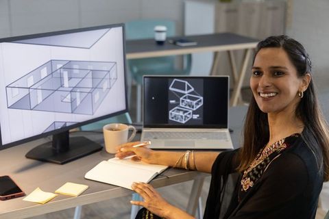 Indian architect designing modern 3d floorplan on dual monitors, smiling at desk workspace
