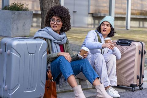 Two friends waiting with luggage and takeaway coffee on city curb, casual travel companions