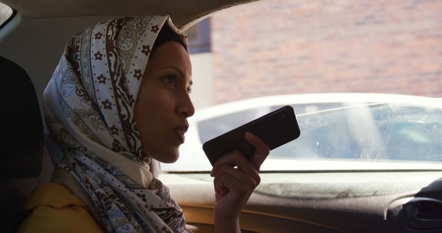 Muslim businesswoman in a car uses a smartphone, with copy space. She's multitasking while commuting, highlighting the blend of modern technology and traditional values.
