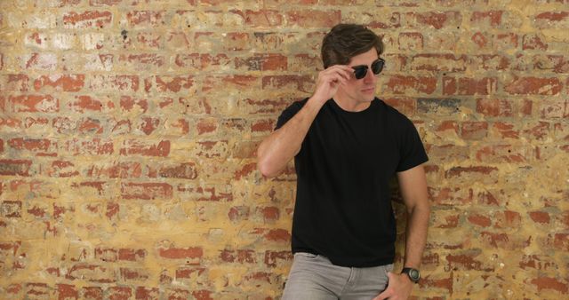 Portrait of happy caucasian man in black t-shirt and sunglasses posing by brick wall. Lifestyle, fashion and style, wellbeing, unaltered.