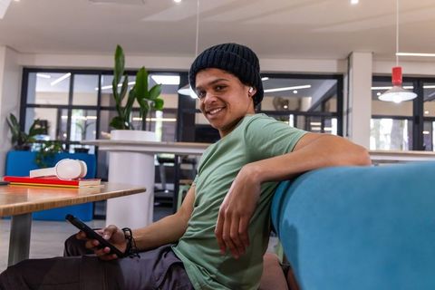 Young man smiling while leaning on blue couch in modern coworking space with smartphone