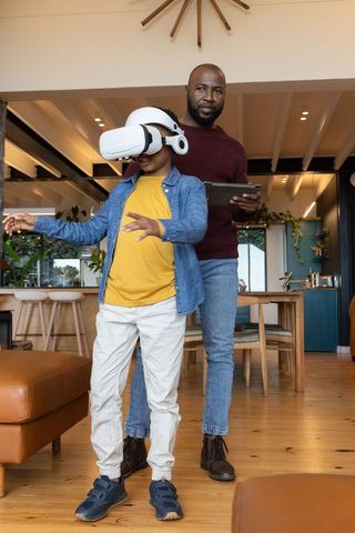 African american father guiding son using vr headset in modern open-plan home while holding tablet