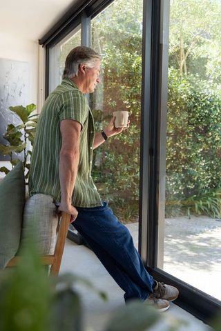 Senior man leaning on sofa by sliding glass doors holding mug and gazing at sunlit garden