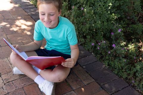 Smiling girl reading on sunlit brick path with red book beside flowering green hedge