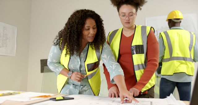 Women architects reviewing building plans together in an office environment, both wearing safety vests and looking at drawings on the table. Background worker wearing a helmet, suggesting an ongoing construction project. Ideal for use in articles related to architecture, construction, teamwork, workplace diversity, and engineering.