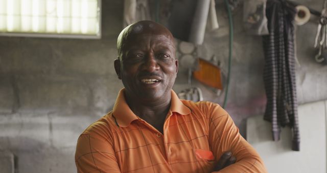 Smiling African American man standing in workshop, arms crossed, and wearing an orange polo shirt. Industrial workspace background featuring tools and repair equipment. Useful for advertisements, articles, and websites focusing on professional skills, industrial jobs, workshops, and craftsmanship.
