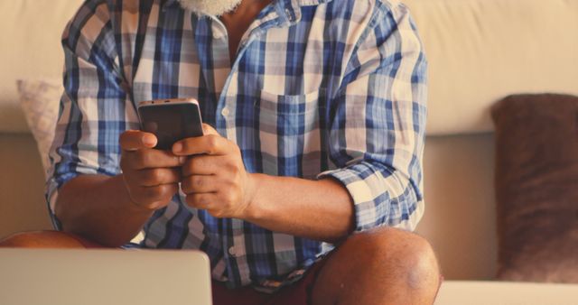 Middle-aged man with beard using smartphone while sitting on couch and wearing plaid shirt, focus on hands. This can be used to illustrate concepts like modern technology, communication, or leisure activities.