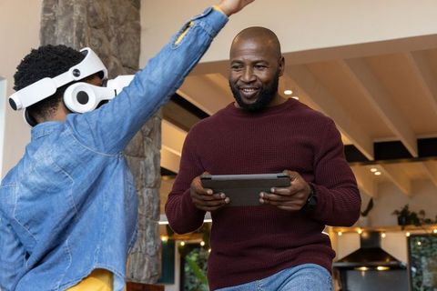 African american man holding tablet while child wearing vr headset playing in kitchen