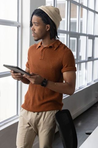 African american man standing by windows holding tablet wearing beige beanie in sunlit studio