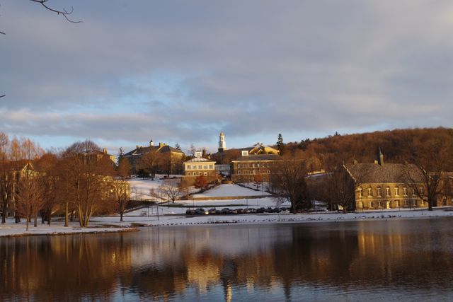 Photo showing a scenic college campus with historic buildings reflecting on a calm lake under an overcast sky. Winter season with snow-covered ground and bare trees adds a tranquil and peaceful ambiance to the scene. Ideal for content about education, university marketing, nature, seasonal reflections, or historic architecture.