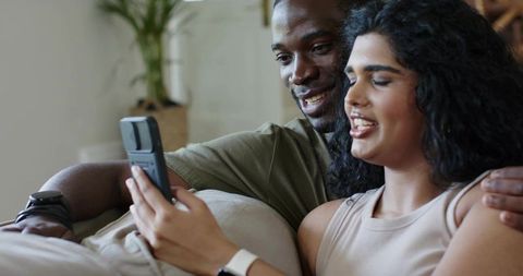 Couple relaxing on sofa sharing smartphone, smiling and scrolling together at home
