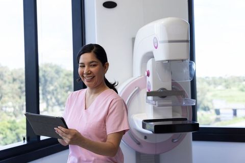 Healthcare worker using tablet next to mammography machine, smiling in radiology clinic