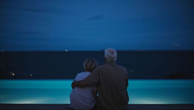 Senior couple embracing at lit infinity pool, watching ocean at night, peaceful companionship