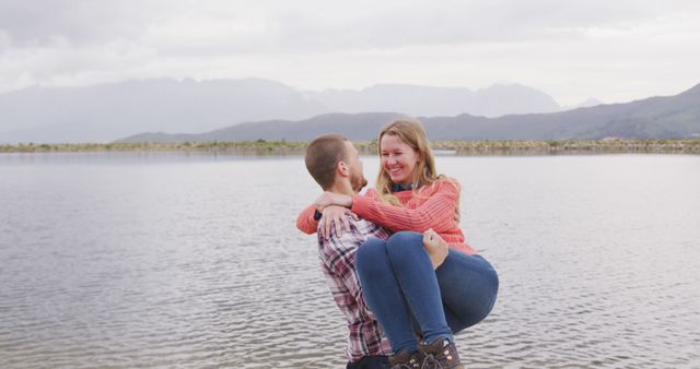 Young couple enjoys playful moment by scenic lake with mountainous backdrop. Ideal for promoting outdoor recreation, romantic getaways, relationship themes, and travel experiences. Perfect for websites, brochures, social media campaigns, and advertisements focusing on couples, travel, and nature retreats.