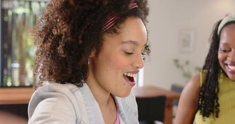 Candid laughing and chatting diverse young women in sunlit kitchen by wooden dining table